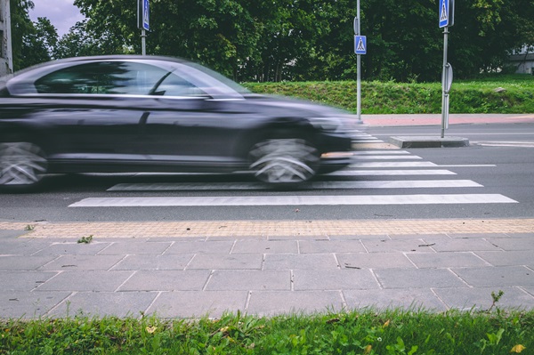A black car speeds through a crosswalk, blurring as it passes over white pedestrian stripes on the road.