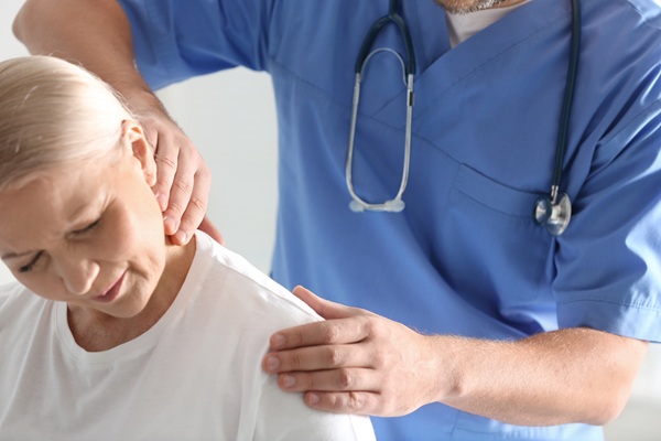 A medical professional in blue scrubs examining the neck and shoulder of an older woman who is wincing in pain.