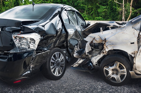 A white car with a crushed front end has collided with the side and rear of a black sedan on a road lined with green trees.