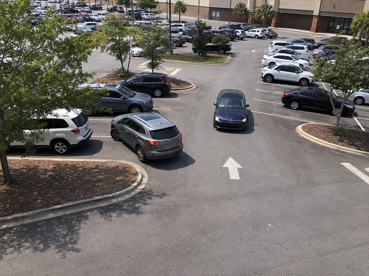 An overhead view of a busy shopping center parking lot with a car navigating a lane between rows of parked vehicles, illustrating the fault questions that arise in Hilton Head and Beaufort parking lot accidents.